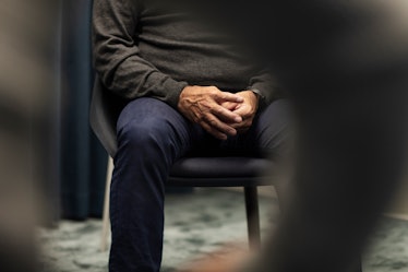 An older man sitting in a chair with his hands resting in front of his groin area