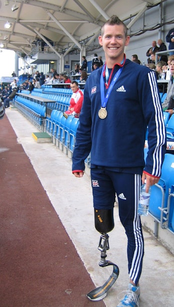 Color photo of a man in athletic wear with a prosthetic leg posing with a gold medal.