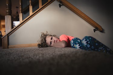 A stubborn child pouting on the carpet near the stairs in her home.