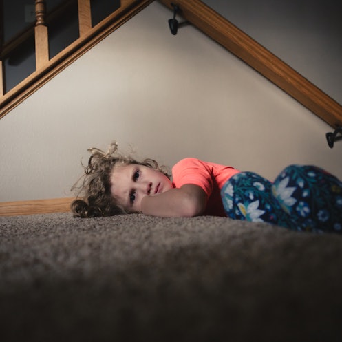A stubborn child pouting on the carpet near the stairs in her home.