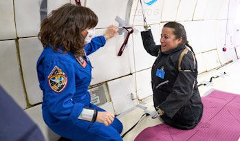Two women, one in a blue flightsuit and one an amputee, talk in the cabin of an aircraft
