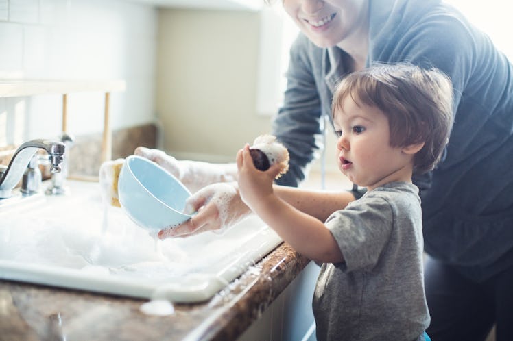 A toddler helping his mom wash dishes in the sink, soap suds are everywhere