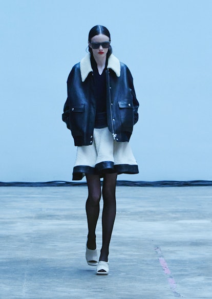 A model wearing a black leather jacket and a white skirt with a black leather edge by Khaite.