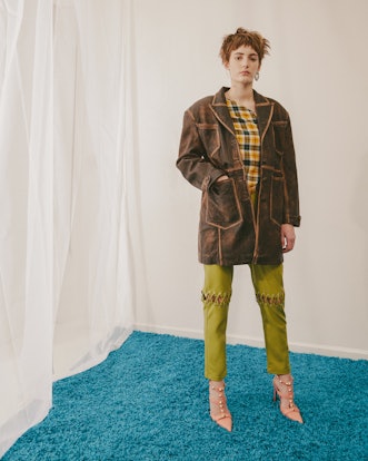 Model for designer Colin LoCascio wearing plaid shirt, a brown coat and green pants with laces on bo...
