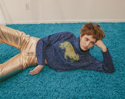 Model for designer Colin LoCascio wearing gold pants and a blue sweater with a green iguana on it