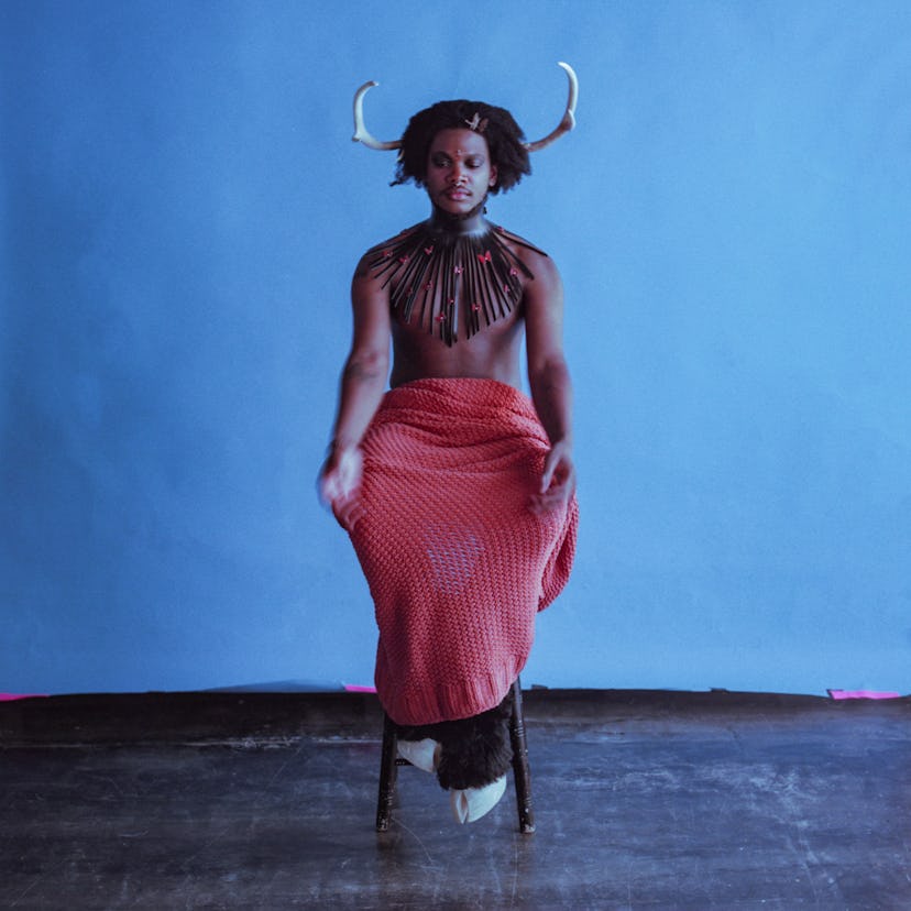 Shamir posing on a stool with two horn accessories, two hoof-shoes and covered with an orange blanke...