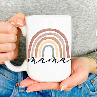Hand holding coffee mug that has a rainbow on it and says "mama"