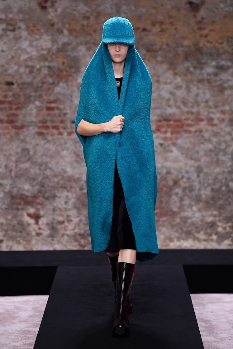 A model in a black Raf Simons dress and blue cape with hat at the London Fashion Week Fall 2022