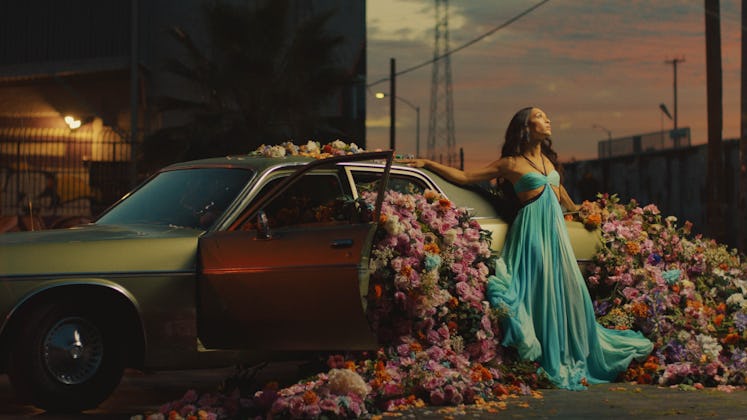 Michaela Jaé leaning against a car that has flowers coming out of it in her video for Something to S...