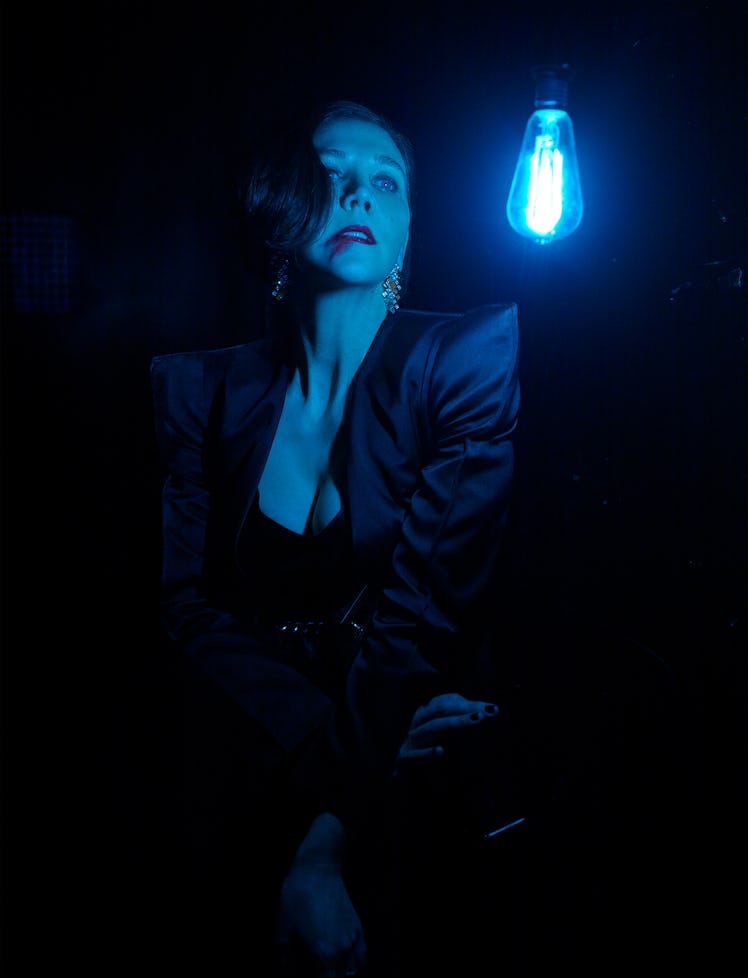 Maggie Gyllenhaal in a black satin blazer looking at a blue glowing lightbulb 