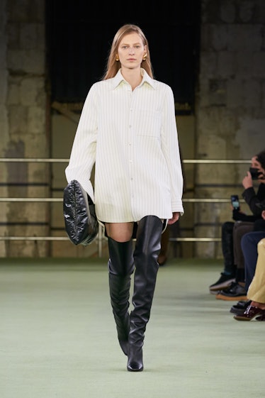 A model in a white oversize shirt and thigh high black leather boots walks at the Bottega Veneta sho...