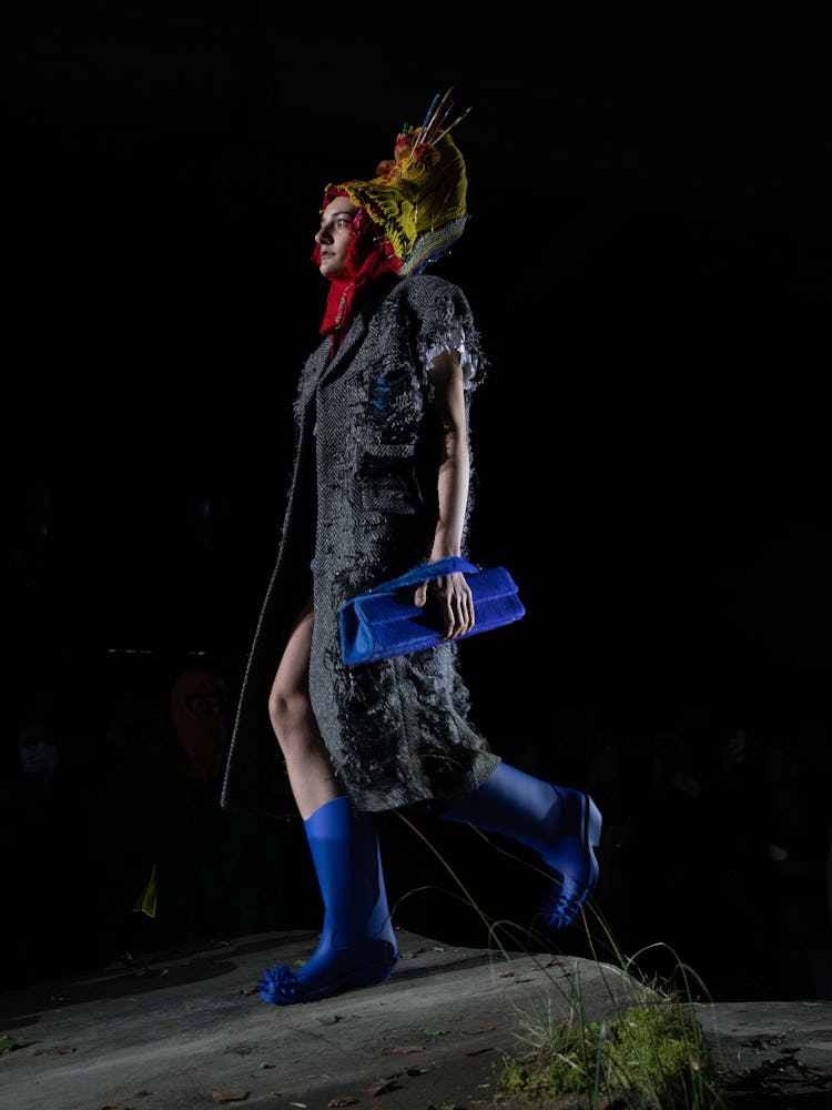 A model in a long grey jacket, blue rain boots and a blue bag walking the Milan Fashion Week runway 