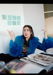 the comedian Catherine Cohen, wearing a blue fur-trimmed coat, throwing her hands up in the air with...