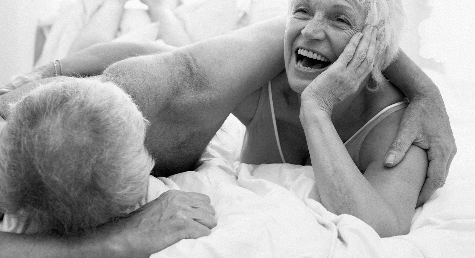 A long-term couple laughing while lying in a bed in their underwear