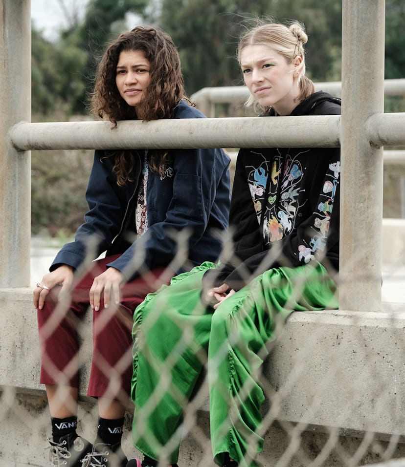 Two women sitting on a concrete barrier with a metal railing, dressed in stylish casual outfits, loo...