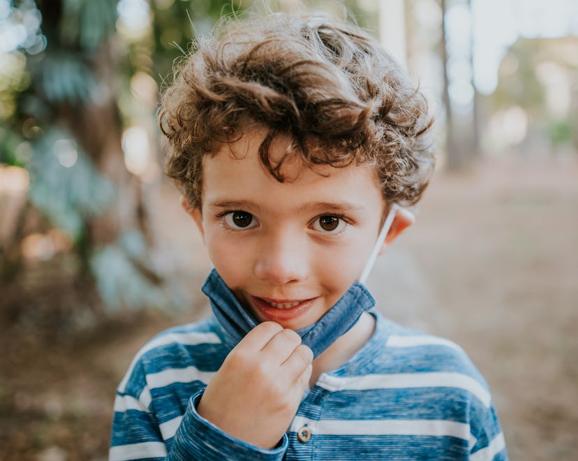 small boy in striped shirt pulls cloth face mask down to reveal smile