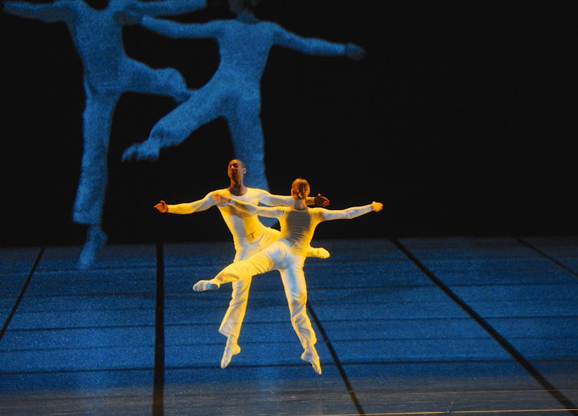 two dancers dressed in all white, leaping across a dramatically lit stage