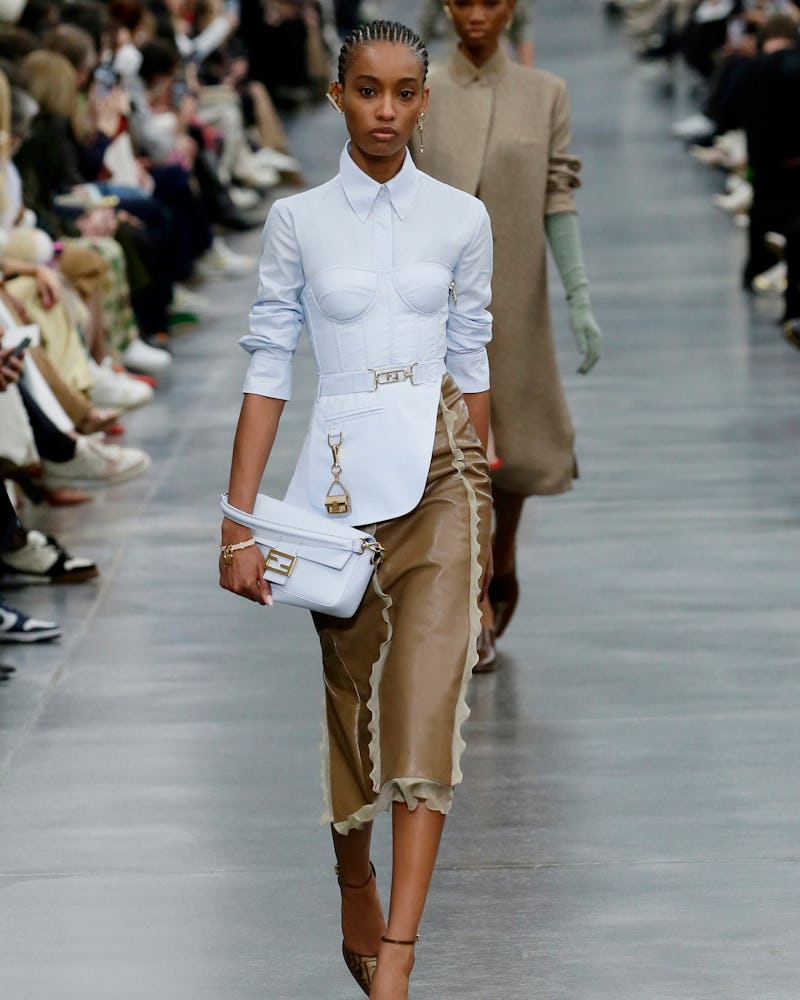 a model wearing a light blue bustier-inspired button-down shirt and brown leather pencil skirt on th...