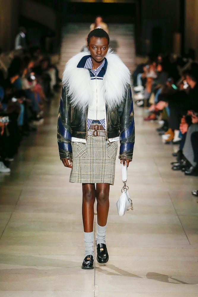a model wearing a plaid skirt, fur-trimmed leather jacket, and polo shirt on the Miu Miu runway
