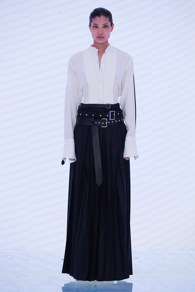 a model wearing white button-down shirt and pleated black maxi skirt with leather buckled belts on t...