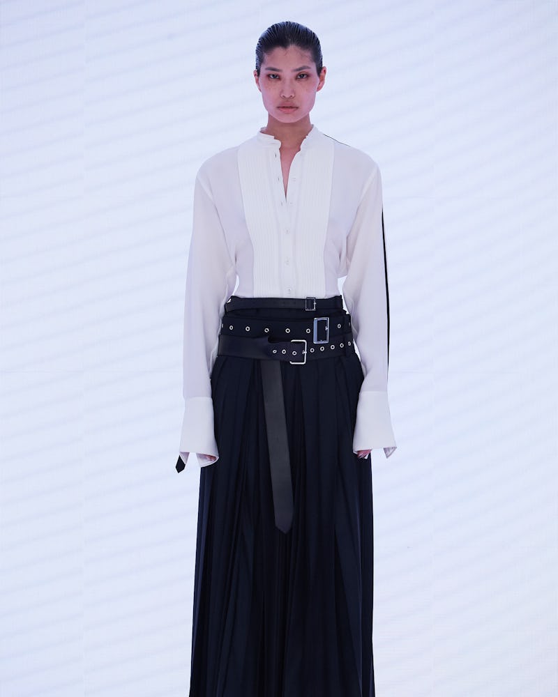 a model wearing white button-down shirt and pleated black maxi skirt with leather buckled belts on t...