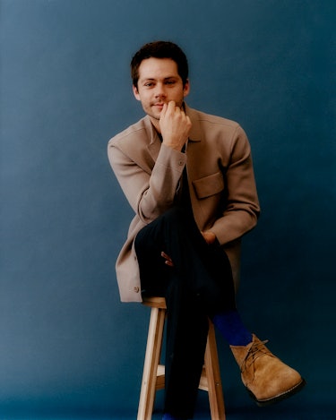 Dylan O'Brien sitting on a high chair in a light brown jacket
