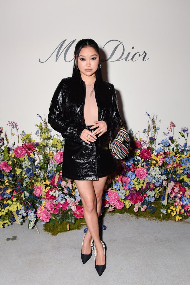 Lana Condor posing in a leather jacket and skirt and a striped purse in front of the floral installa...