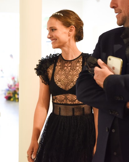 Natalie Portman smiling in a black lace and tulle dress with flower embroidery on the top, and a gol...
