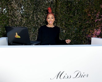 DJ Kitty Ca$h smiling behind the counter with the "Miss Dior" sign and her laptop.