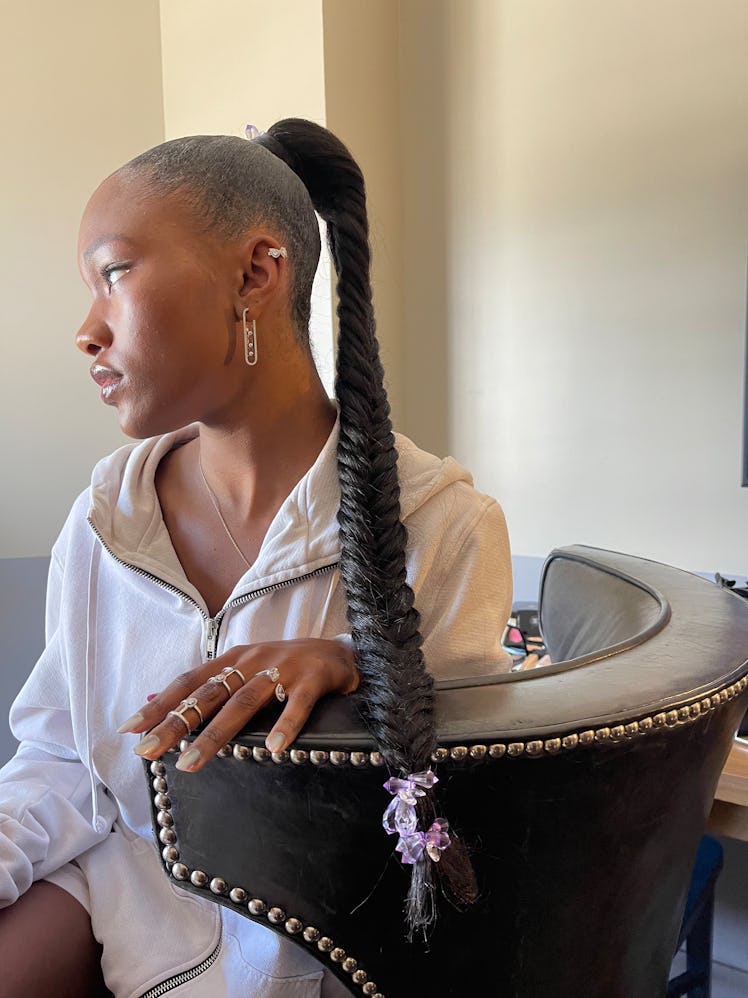 A side-profile of Demi Singleton  with a long braid in a white hoodie sitting and looking into the d...