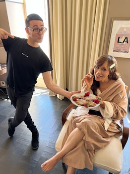 Christian Siriano and Rosie Perez in a beige house cape holding a plate with food while getting read...
