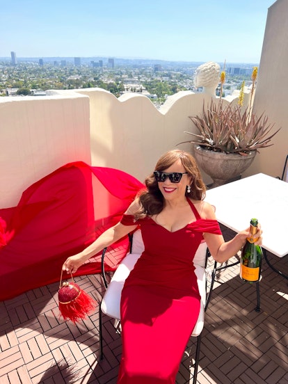Rosie Perez in a red gown, holding a bottle of champagne while sitting on a lounge chair on a terrac...