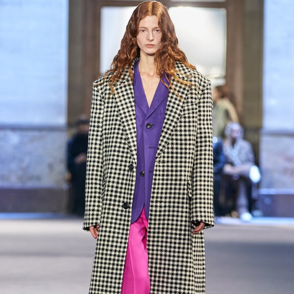 a model wearing a houndstooth check coat, a purple blouse, and pink pants on the Ami runway