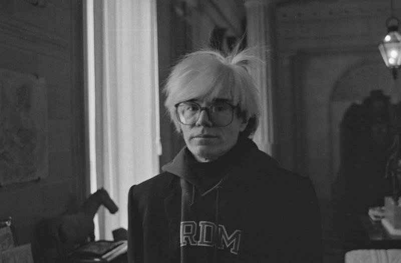 An old black and white photograph of Andy Warhoo. He's wearing glasses, a hoodie, and a blazer and s...