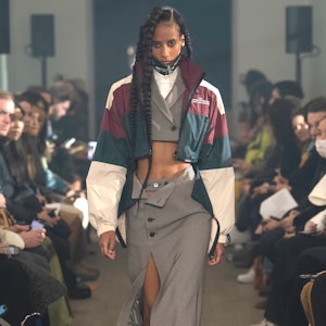 a model wearing a cropped blazer and pencil skirt with a varsity windbreaker jacket on the Rokh runw...