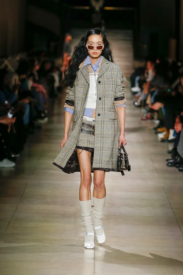 a model wearing a plaid miniskirt and coat with a striped button down and cropped sweater on the Miu...