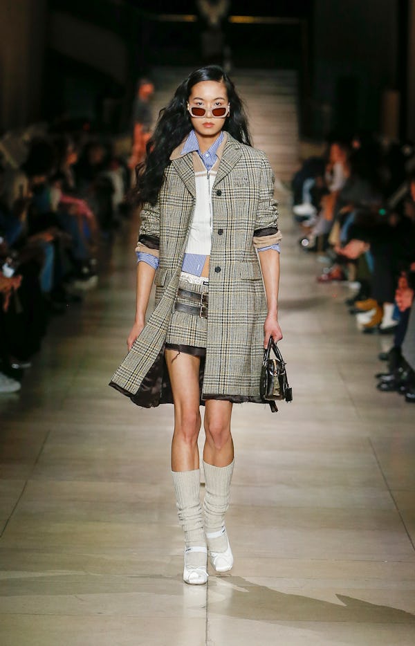 a model wearing a plaid miniskirt and coat with a striped button down and cropped sweater on the Miu...
