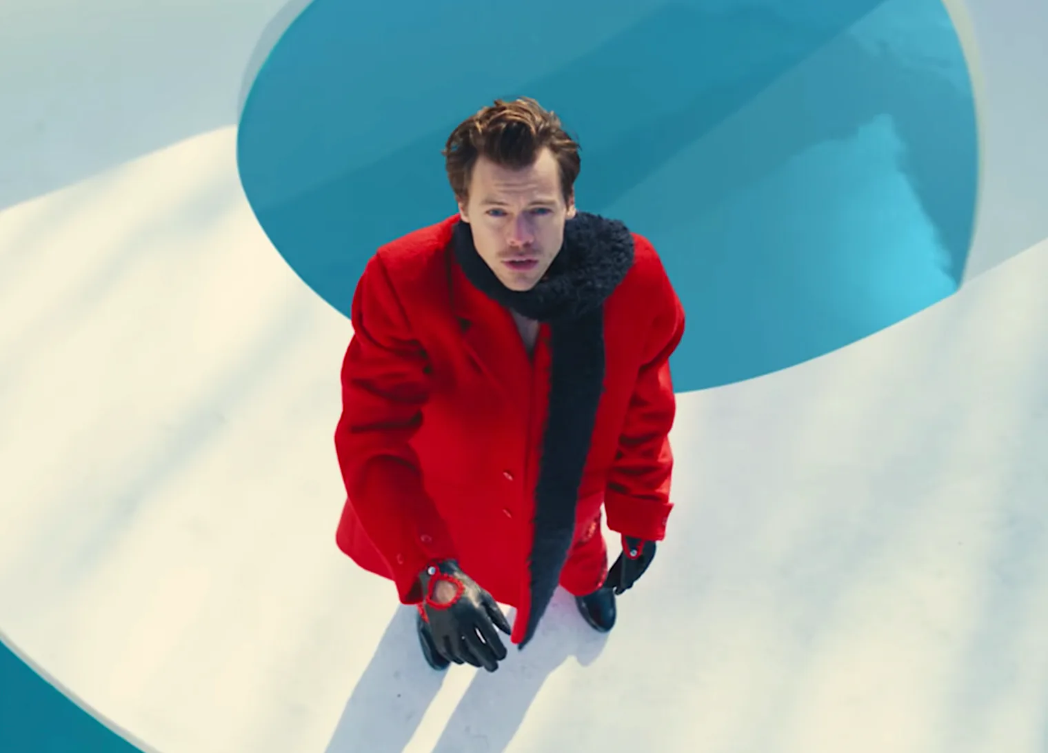Harry Styles, wearing a red coat and black scarf and gloves, looking up in his 'As It Was' music vid...