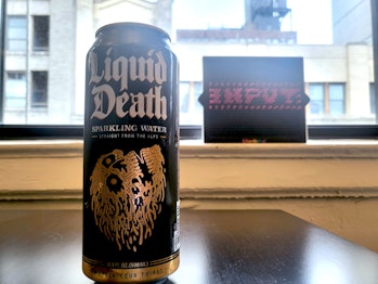 A black and gold can of Liquid Death sparkling water with the Input logo on a lite-brite in the back...