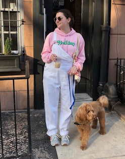 A woman in a pink hoodie, white overalls and a pair of staple sneakers with her dog leaving in front...