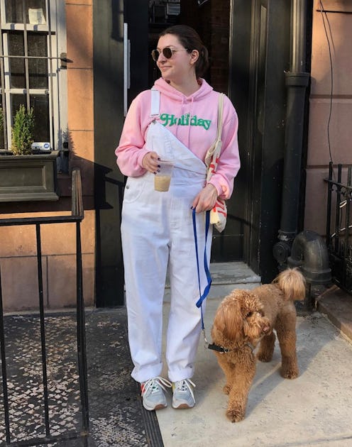 A woman in a pink hoodie, white overalls and a pair of staple sneakers with her dog leaving in front...