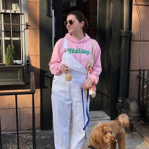 A woman in a pink hoodie, white overalls and a pair of staple sneakers with her dog leaving in front...
