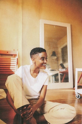 Syd sitting on the floor in a white tee and beige pants, looking to the side with a smile