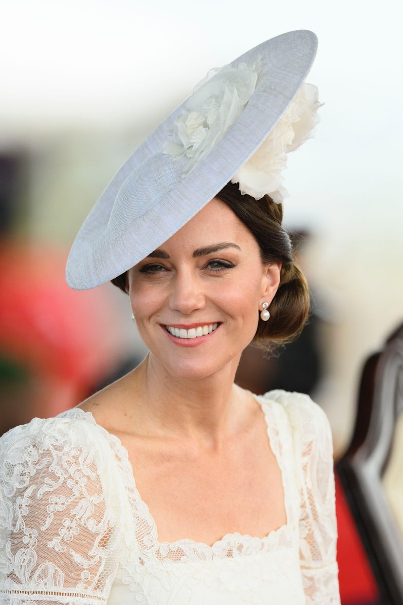 Kate Middleton wears a white flat disc-shaped hat tilted to the side of her face with a low chignon ...