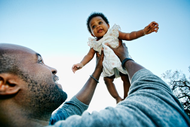 A Black father holding his baby girl high against a blue sky with the sun shining behind her. The gi...
