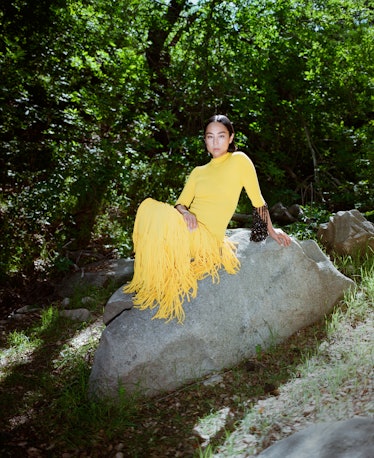 Greta Lee in a yellow fringed Proenza Schouler dress sitting on a rock