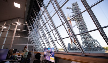 A person looks out of a large window, through which a rocket and a tall metal structure are seen.