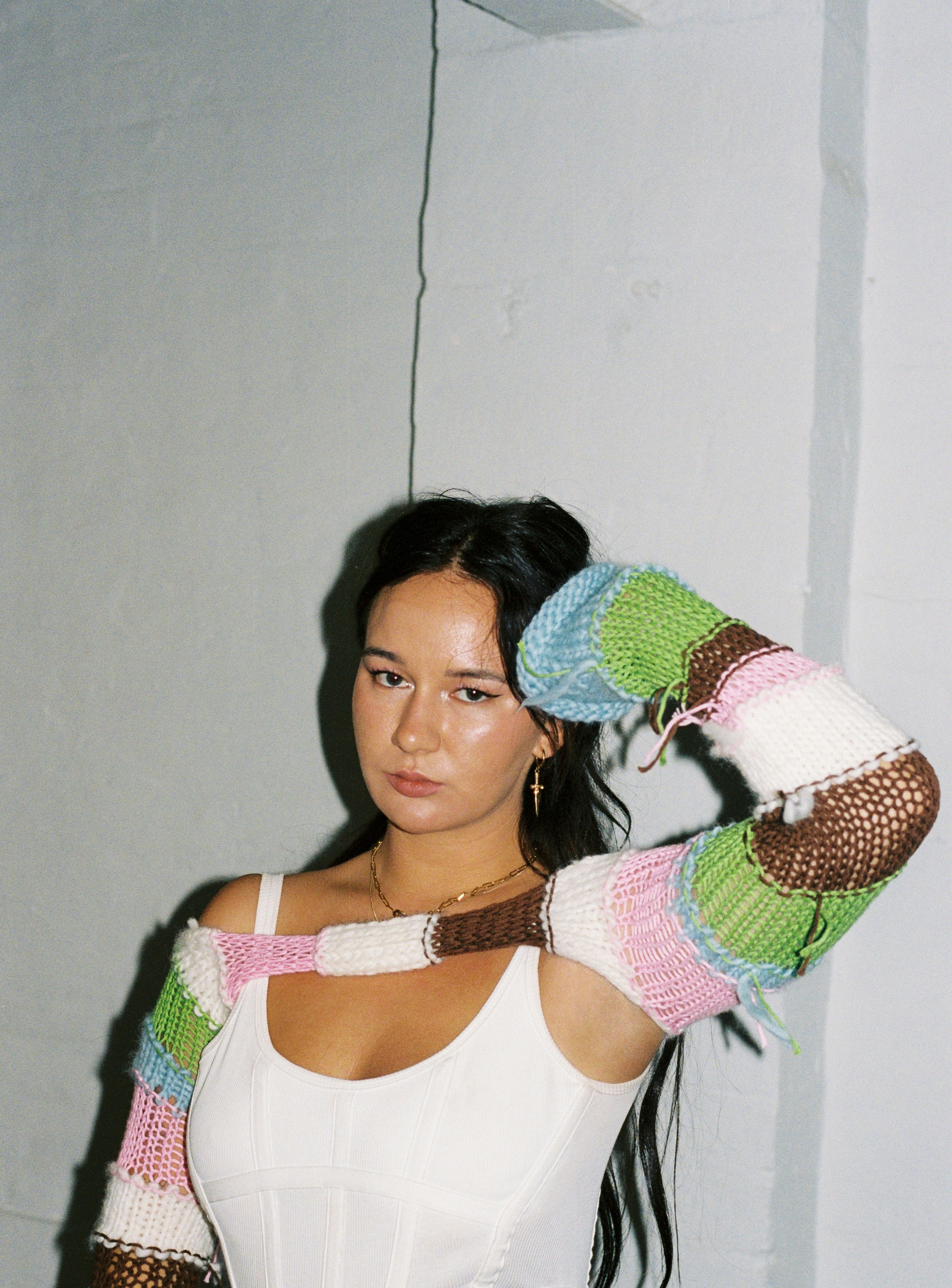 Australian musician Mallrat posing in a white undershirt and a torn white, brown, green, pink, and b...