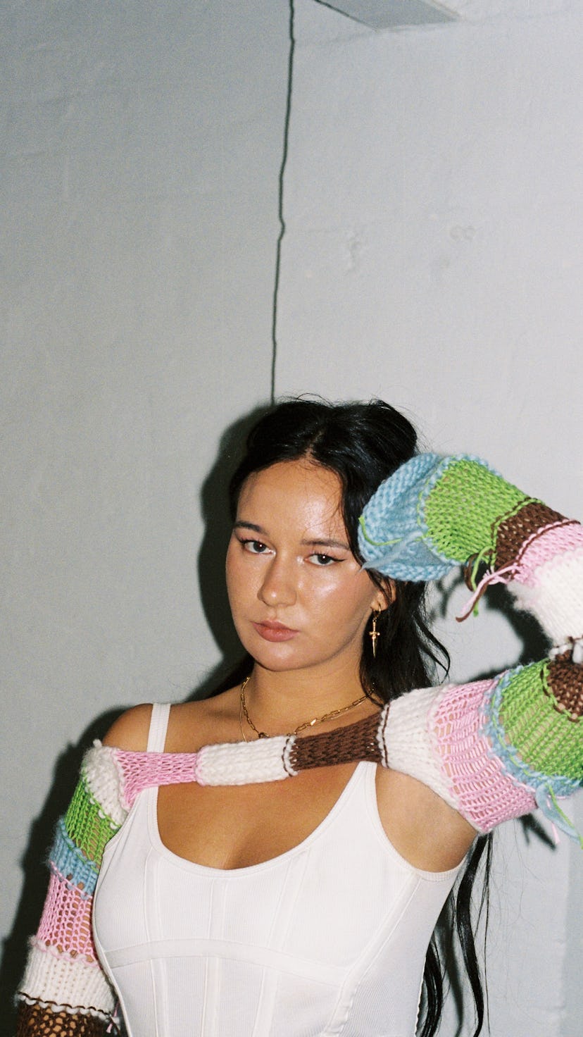 Australian musician Mallrat posing in a white undershirt and a torn white, brown, green, pink, and b...