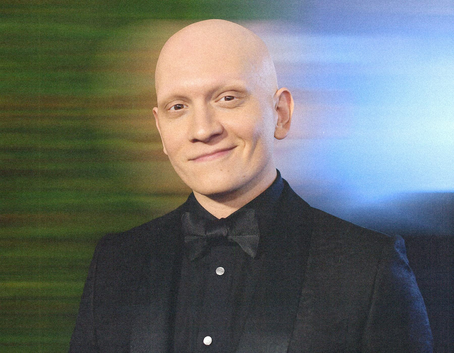 Anthony Carrigan wearing a black suit and smirking at the camera